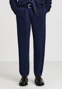 Uomo che indossa pantaloni blu navy con una cintura abbinata, scarpe eleganti in cuoio nere e un maglione lavorato a maglia blu navy con le mani in tasca.