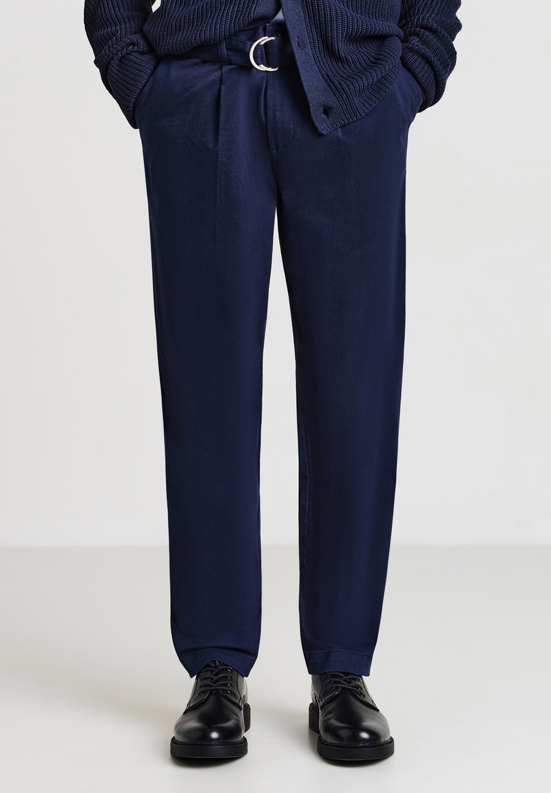Uomo che indossa pantaloni blu navy con una cintura abbinata, scarpe eleganti in cuoio nere e un maglione lavorato a maglia blu navy con le mani in tasca.