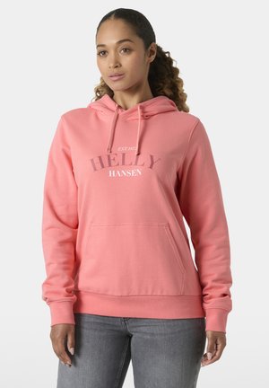 Vrouw met krullend haar, gekleed in een roze Helly Hansen hoodie en grijze jeans, staand tegen een effen lichte achtergrond.