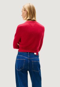 Roter Pullover mit geripptem Saum und V-förmigem Ausschnitt, getragen über blauen, hoch taillierten Jeans mit Gesäßtaschen und einem Etikett.