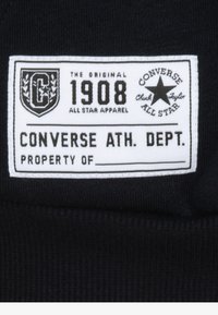 Converse címke fekete háttérrel, fehér szöveggel és díszes emblémával. A szöveg tartalmazza: "AZ EREDETI", "1908" és "CONVERSE ATH. DEPT."
