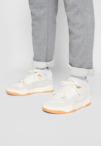Zapatillas blancas con acentos naranjas, materiales de malla y ante, cuello acolchado y suela texturizada, combinadas con pantalones de chándal grises con puños.