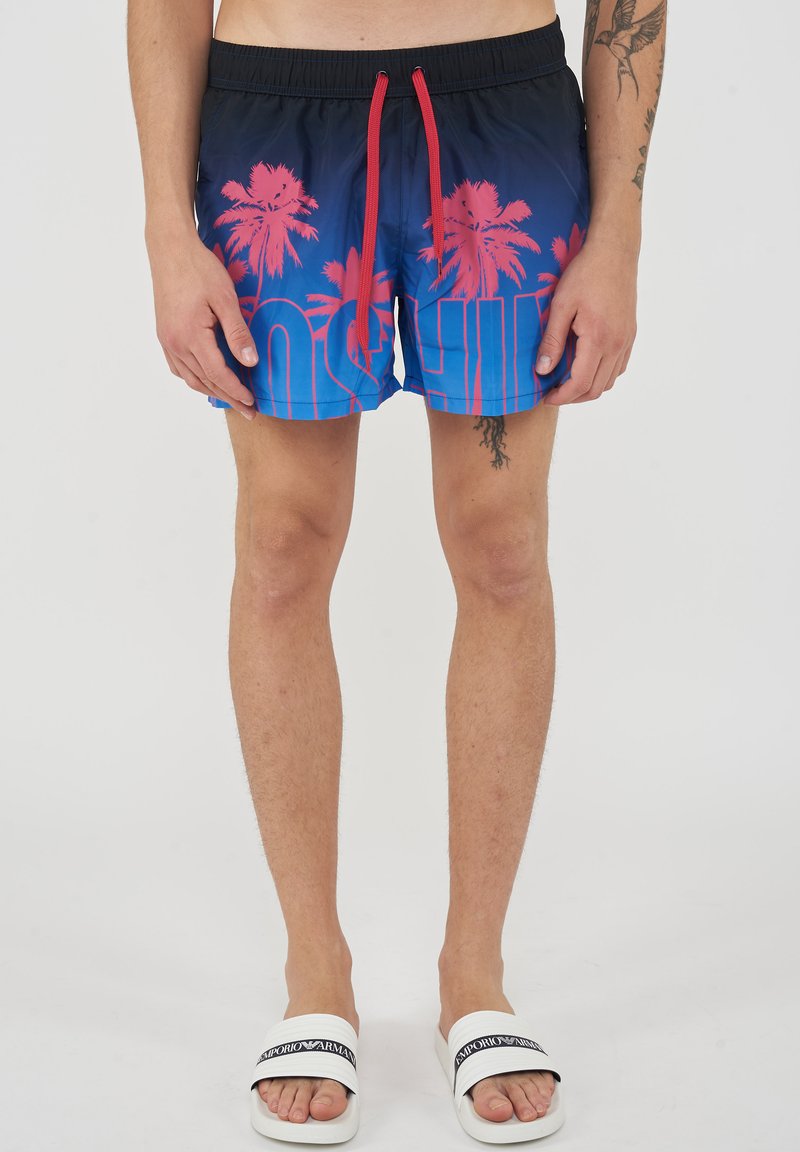 Men's swim shorts with red palm tree print and red drawstrings, worn with white Emporio Armani slides on light skin legs.