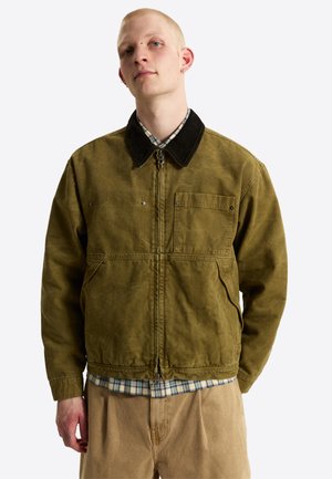 Jonge man draagt een olijfgroene jas met rits en zwarte kraag over een geruit overhemd en beige broek, staand tegen een effen achtergrond.
