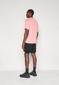 Κοντομάνικο, μπλουζάκι με στρογγυλή λαιμόκοψη σε ανοιχτό ροζ ύφασμα, σε συνδυασμό με μαύρο σορτς. Το μοντέλο φορά μαύρα αθλητικά παπούτσια και κάλτσες. Πλάγια όψη από πίσω.