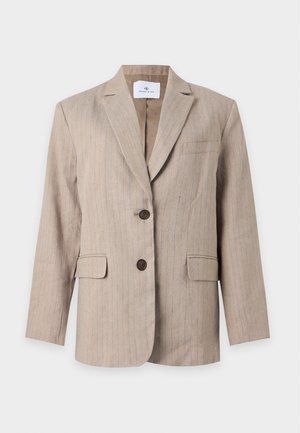 Femme debout, souriante, portant un blazer beige avec short assorti, un haut blanc et des ballerines noires sur un fond uni clair.