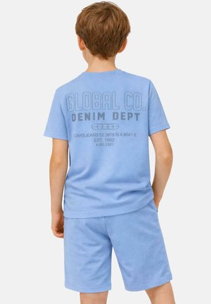 Jongen met kort bruin haar, draagt een lichtblauw T-shirt en korte broek met de opdruk "GLOBAL CO. DENIM DEPT" op de achterkant.