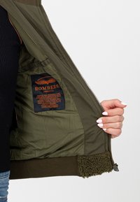 Oliwkowa kurtka bomberka z zapięciem na zamek, puszyste brązowe podszycie i niebieska metka wewnątrz z logo marki "BOMBERS ORIGINAL."