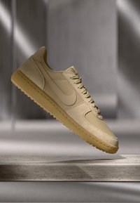 Beige sport sneaker met gestructureerde zool, veters en swoosh-logo, zwevend boven een stenen oppervlak in zachte verlichting.