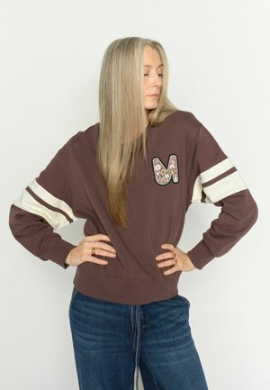 Vrouw met lang blond haar die een bruine trui draagt met witte strepen op de mouwen en een bloemige "M"-patch, poserend met handen in de zij.