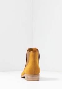 Stivaletto alla caviglia in camoscio giallo con pannelli elastici laterali, tirante posteriore e tacco basso impilato, che presenta una texture liscia e un design minimalista.