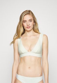 Bralette texturizado verde claro com um decote em V profundo e alças largas. As parte de baixo do biquíni correspondente são visíveis. Design liso e minimalista.