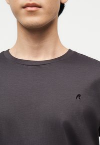 Κοντινό πλάνο ενός ατόμου που φορεί σκούρο γκρι T-shirt με στρογγυλή λαιμόκοψη και μικρό μαύρο κεντημένο λογότυπο "R" στο αριστερό στήθος.