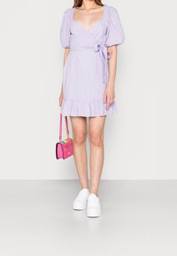 Lavendel kort omslagklänning med puffärmar, volangkant och ögla detaljer. Kombinerad med en ljusrosa handväska och vita sneakers.