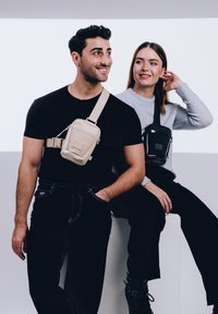 Két modell bemutatja a minimalista crossbody táskákat: egy világos bézs táska a férfin, és egy fekete táska a nőn, mindkettő állítható pánttal és cipzárral.