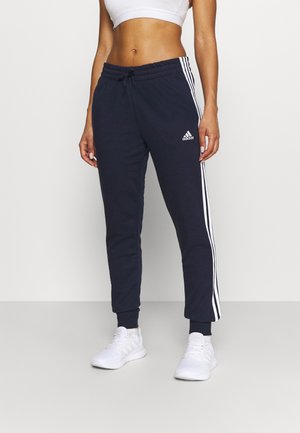 Personne en forme portant un pantalon de survêtement Adidas bleu marine avec des bandes blanches, des baskets blanches et un crop top de sport blanc, debout devant un fond uni.