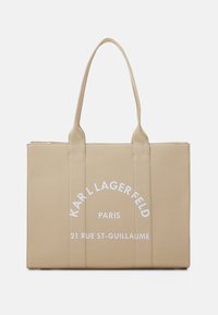 Tan canvas-kassi, jossa on kaksinkertaiset kahvat ja valkoista painettua tekstiä: "KARL LAGERFELD PARIS 21 RUE ST-GUILLAUME." Suorakaiteen muotoinen.