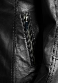 Detalj crne kožne jakne s glatkom teksturom, otkrivenim patentnim zatvaračem u bočnoj džepu i ušivenim šavovima duž rubova.