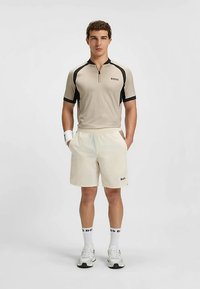 Mannen sportieve outfit met een lichtbeige polo met korte mouwen, zwarte accenten en logo, gecombineerd met witte korte broek en witte sportschoenen.