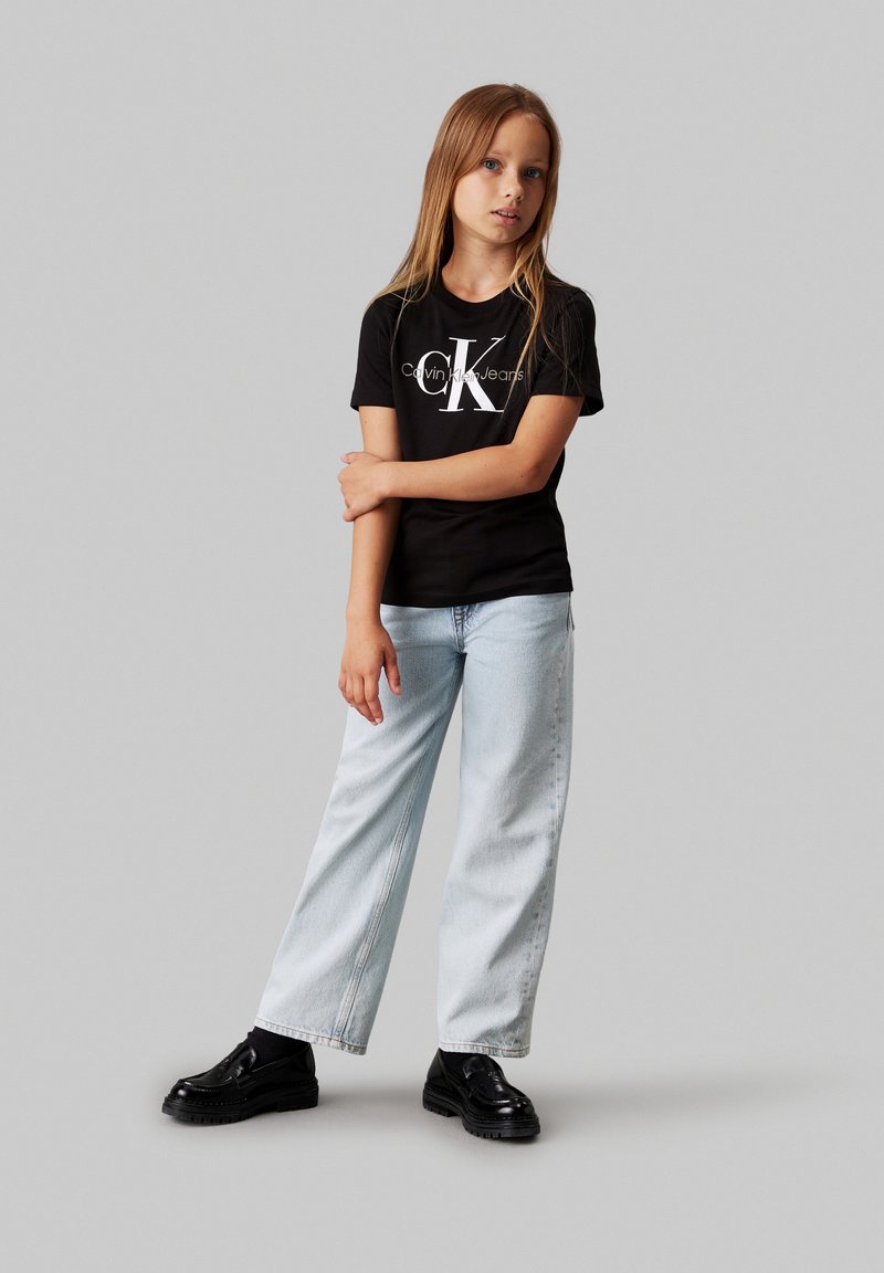 Melna īsām piedurknēm T-kreklu ar baltu "Calvin Klein Jeans" logo, apvienots ar gaiši ziliem platajiem džinsiem un melniem masīviem apaviem.