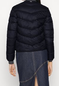 Žena v černé prošívané bundě s šikmým prošíváním a tmavých džínech se zadním rozparkem, zachycena zezadu.