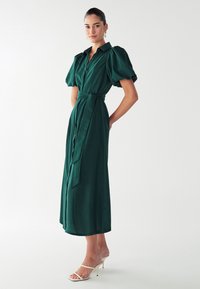 Zelena obleka s kratkimi napihnjenimi rokavi, pasom in krilom srednje dolžine. Gladka tekstura in tekoč dizajn, v kombinaciji z belimi sandalmi na visoki peti.