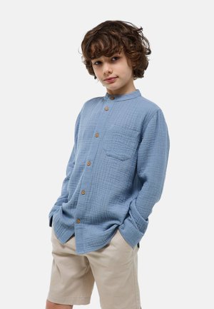 Un garçon aux cheveux bruns bouclés se tient les mains dans les poches, portant une chemise bleue boutonnée et un short beige, sur un fond uni.