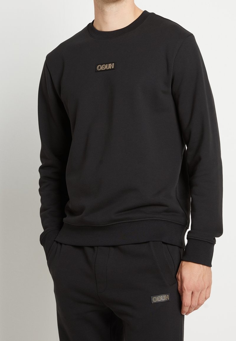 Homme portant un sweat-shirt noir à manches longues et un pantalon noir assorti, tous deux avec de petits logos rectangulaires portant l'inscription « HUGO » en beige.
