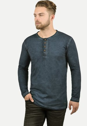 Majica s dugim rukavima u tamnoplavoj boji, model henley, s okruglim izrezom, placket-om s pet dugmadi i istrošenom teksturom, uparenim s tamnim trapericama.