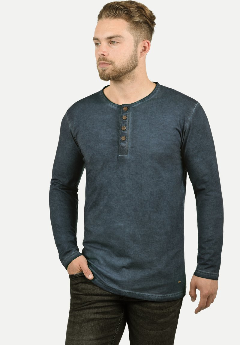 Langermet, marineblå henley-skjorte med rund hals, fem knappelukkinger og en slitt tekstur, kombinert med mørke jeans.