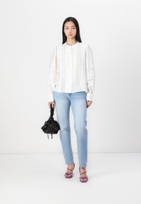 Witte blouse met knoopsluiting, verticale plooien en kantdetails, lichtblauwe spijkerbroek, zwarte geprinte tas en paarse sandaaltjes met bandjes.