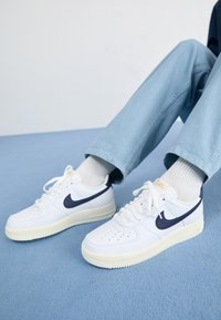 Tênis brancos de couro com o símbolo da Nike em azul-marinho, biqueira perfurada e sola de borracha creme. Combinados com calças azul claro e meias brancas.