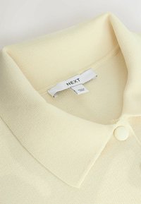 Pull jaune clair à col avec tissu texturé, comportant un bouton rond et une étiquette portant la mention "NEXT" attachée à l'encolure.