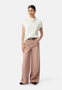 Camiseta blanca de manga corta combinada con pantalones de pierna ancha ligeros de color rosa claro y un cinturón marrón. La modelo lleva zapatos oscuros y collares de oro en capas.