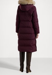 Dlouhá pufová bunda v barvě švestkové s kapucí podšitou kožešinou, obsahující horizontální prošívané části a boční švy pro lepší pohyb.