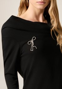 Zwart topje met een brede halslijn en lange mouwen, voorzien van een strikbroche met strasstenen aan de linkerkant, die een decoratief detail toevoegt.