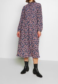 Långärmad klänning med marinblå bakgrund och rosa blommig paisleymönster, hög halsringning och volangkant, kombinerad med svarta ankelskor.