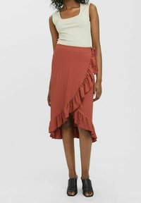 Spódnica w kolorze rdzy z falbaniastym dołem, wykonana z teksturowanej tkaniny, zestawiona z jasną bluzką na ramiączkach i czarnymi sandałami odkrytymi na palcach.