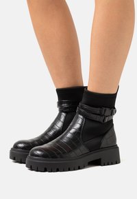 Bottines noires avec dessus synthétique à motif crocodile, côtés élastiques et semelle en caoutchouc épaisse. Comprend des accents de lanières ajustables.