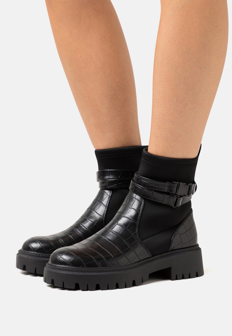 Bottines noires avec dessus synthétique à motif crocodile, côtés élastiques et semelle en caoutchouc épaisse. Comprend des accents de lanières ajustables.