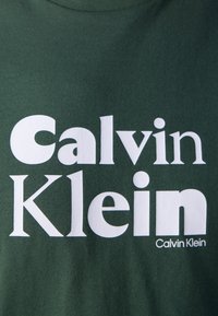 White "Calvin Klein" logo text prominently displayed on dark green fabric background.