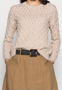 Béžový pletený svetr s vzorem copánků, kulatým výstřihem, dlouhými rukávy a žebrovanými manžetami. Nositelný přes hnědou sukní s černým texturovaným páskem.