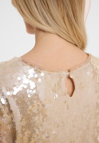 Top en sequins dorés avec une texture en mesh doux, un col rond et un détail en goutte d'eau au dos. Les sequins créent un motif réfléchissant.