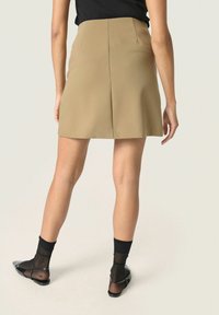 Khaki mini szoknya egyenes szabással, sima textúrával és hátsó redőzött részlettel. Fekete áttetsző zoknikkal és lapos cipőkkel párosítva.