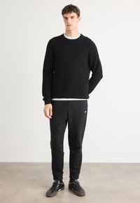 Svart stickad tröja med långa ärmar, tillsammans med svarta byxor med en diskret logotyp. Avslappnade skor fulländar outfiten.