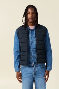 Chaleco acolchado negro sobre una chaqueta de mezclilla azul. El chaleco presenta secciones acolchadas. El conjunto incluye jeans de mezclilla azules y una camisa rosa clara.