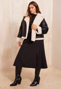 Svart faux läder kort jacka med krämfärgad shearling-krage och manschetter, tillsammans med en lång svart klänning med sidssprund och knähöga svarta stövlar.