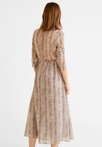 Pitkä, läpikuultava beige mekko, jossa on käärmeennahkakuosi. Mekossa on pitkät hihat, vyötärö on kootusti, ja laahus on virtaava ja ulottuu polvien alapuolelle.
