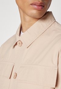 Chaqueta beige de botones con un tejido texturizado, un cuello de punta y dos bolsillos en el pecho, asegurados con botones de color claro.