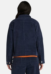 Marinblå kordjacka med krage, ryggparti och tryckknappdetaljer. Bärs över en orange, strukturell tröja och har en boxig passform.